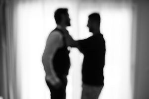 Two silhouetted figures stand in conversation against a bright background in a dramatic black and white composition.