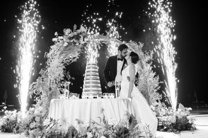 Newlyweds stand by their wedding cake with sparklers creating dramatic lighting effects under a decorated arch.
