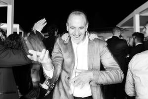 A person in a light colored suit smiles and gestures while standing among a crowd at an evening event.