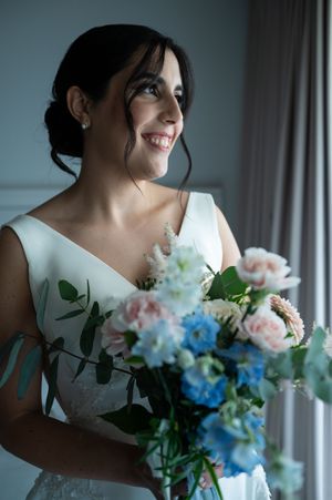 A bride in a white dress holds a romantic bouquet of pink and blue flowers against a soft window light.