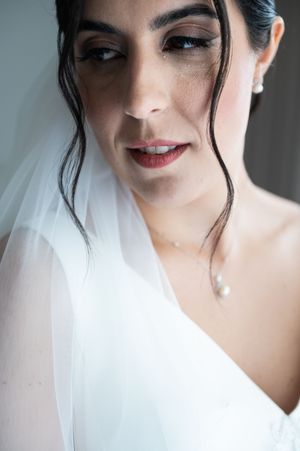 Close-up portrait of a bride wearing a white veil and pearl necklace with red lipstick and dark hair.