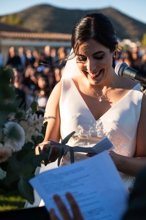 Person in white dress looking down and reading from paper at outdoor event with blurred background.