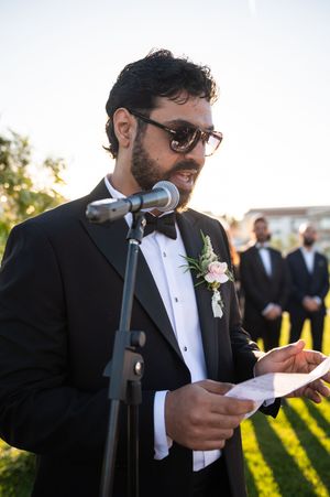 A person in a black tuxedo and sunglasses speaks at a microphone during an outdoor wedding ceremony.
