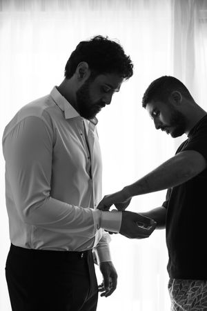 Black and white silhouette of two men in formal wear, with one adjusting the other's sleeve cuff in a pre-event moment.