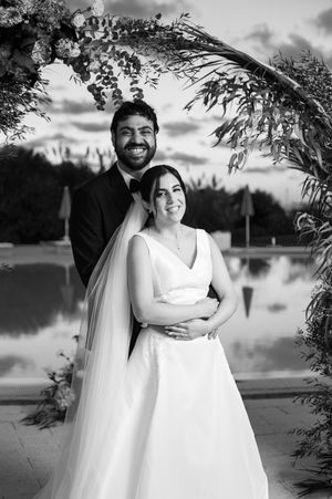 Black and white wedding portrait of a couple embracing under a floral arch by a lake at sunset.