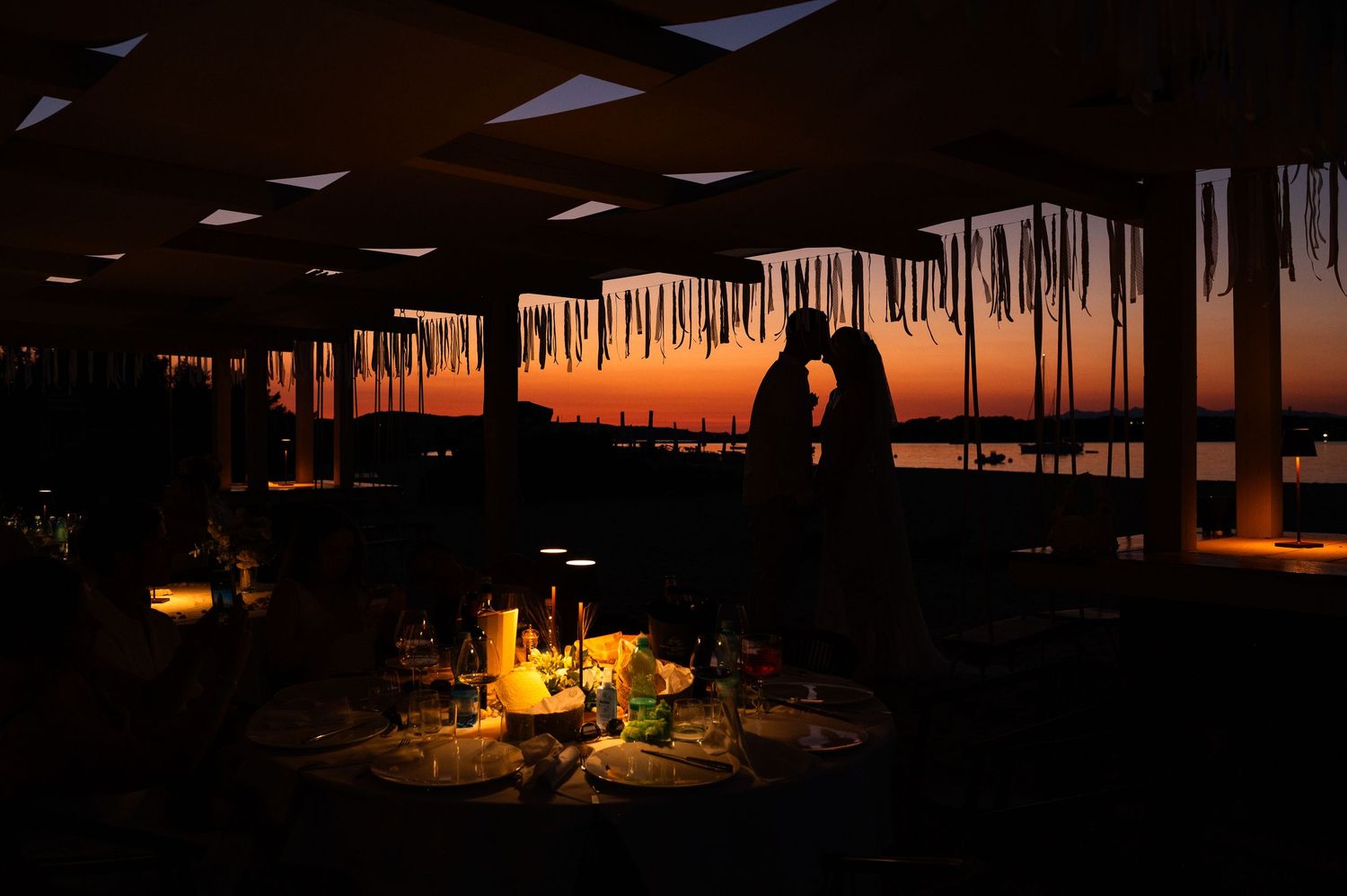 Romantic silhouette of a couple kissing at sunset on a decorated outdoor terrace with candlelit tables.