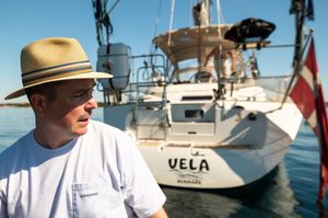 Man in straw hat standing near sailboat named Vela docked at marina on sunny day.