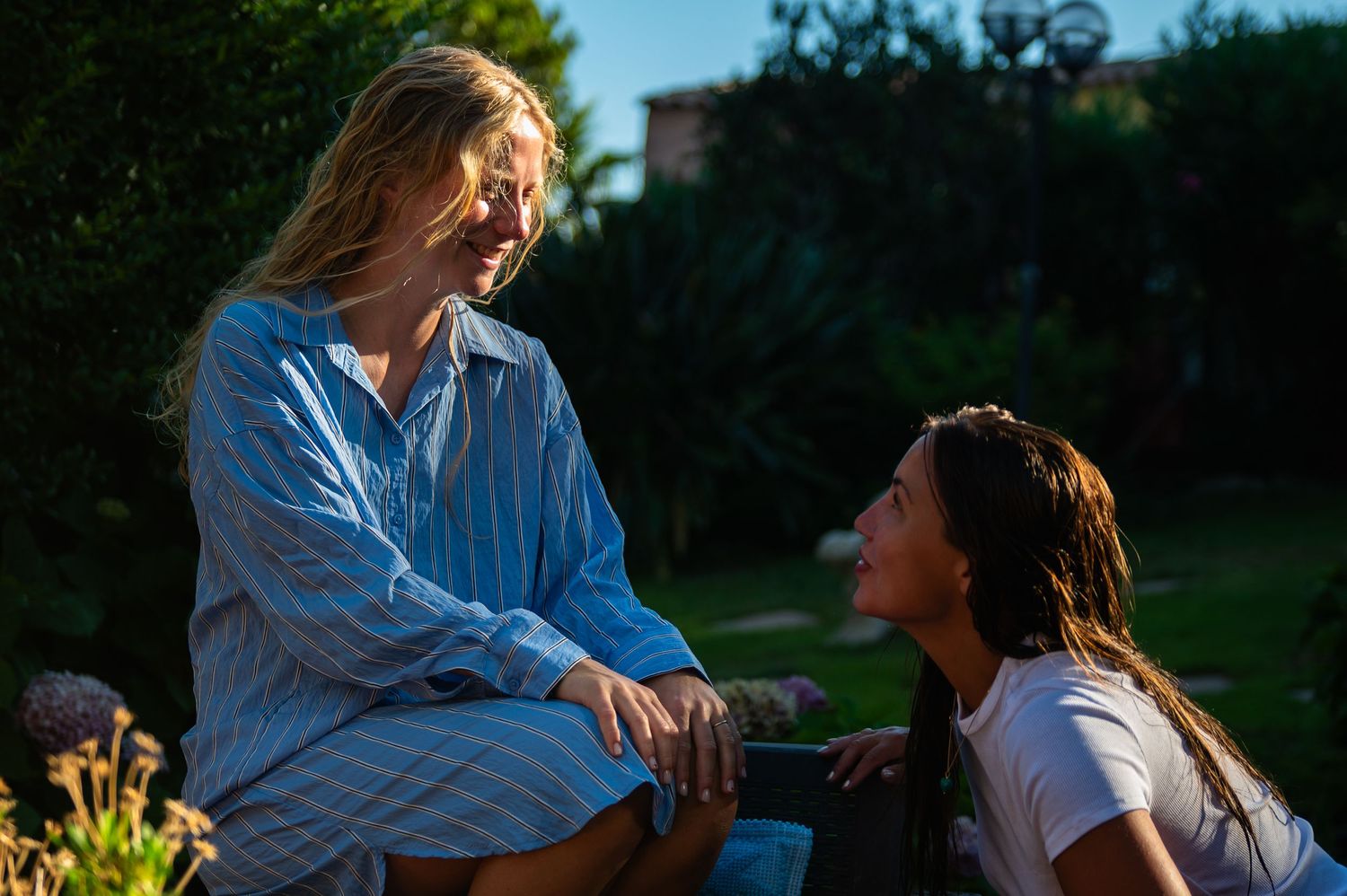 Two women sharing a joyful conversation outdoors in a sunlit garden, one in a blue striped shirt.
