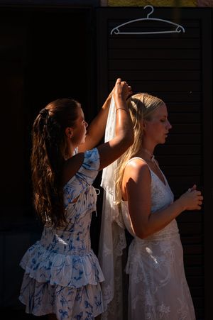 Bridesmaid in floral dress adjusting veil on blonde bride in white gown outdoors in golden sunlight.