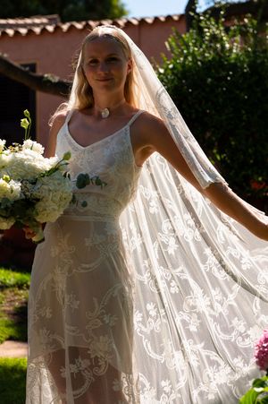 Blonde bride in lace wedding dress holding white bouquet, wearing veil outdoors in sunlit garden.