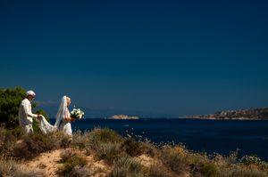Bride and groom in white standing on rocky coastal cliffside overlooking deep blue Mediterranean sea on sunny wedding day.