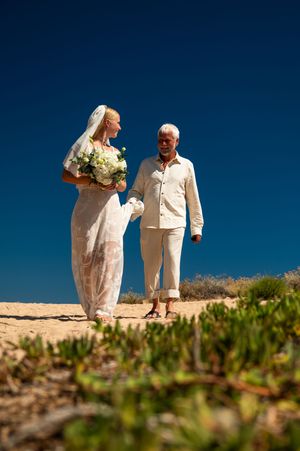 Bride in white gown holding bouquet walks barefoot on sandy beach with older man under clear blue sky.