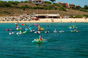 Colorful inflatable floats and swimmers enjoy crystal-clear turquoise waters near a sandy beach resort on a sunny day.