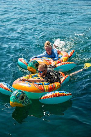 Two people riding colorful inflatable turtle floats on bright turquoise ocean water on a sunny day.