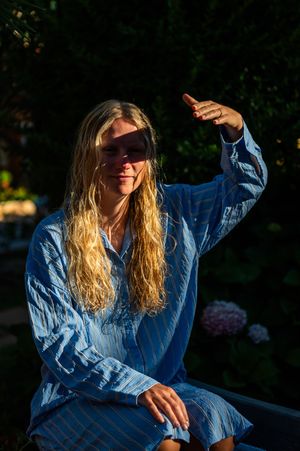 Woman with long blonde hair in blue striped dress sitting outdoors, shielding eyes from sunlight in garden.