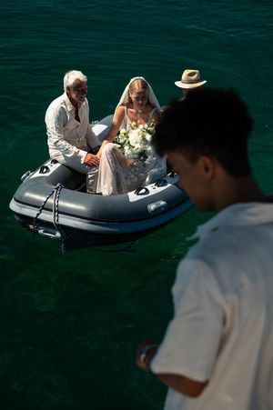 Bride in veil with bouquet and older man arrive by inflatable boat on turquoise water as guests watch from dock.