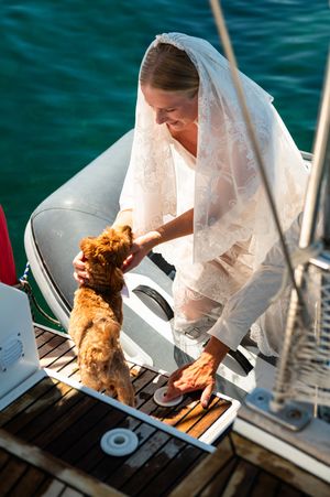 Bride in white lace veil petting a fluffy brown dog on a sailboat deck with turquoise water behind.