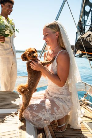 Bride in lace wedding dress and veil playing with brown poodle on sailboat deck during outdoor wedding ceremony.