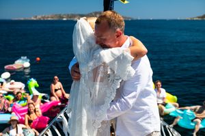 Couple sharing emotional wedding hug on boat deck with colorful floaties and guests in background.