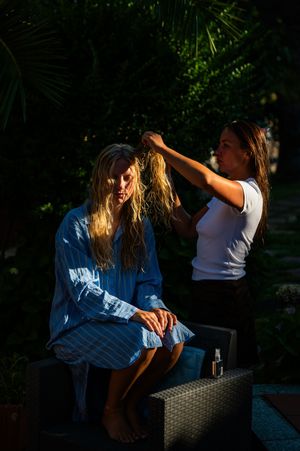 Woman styling blonde hair outdoors in sunlight, surrounded by lush green tropical plants.