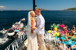 Bride and groom share a kiss on a boat deck surrounded by guests on colorful floaties in the sparkling blue sea.