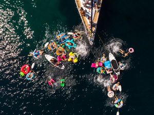 Aerial view of a large group of people swimming and floating on colorful inflatables beside a sailboat in dark blue water.