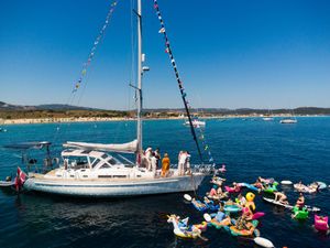 People enjoying a sunny day on a sailboat with colorful flags, surrounded by inflatable floats in calm blue waters.