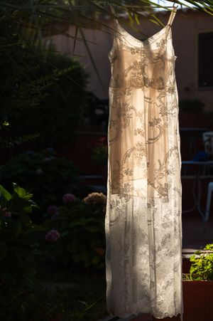Elegant cream lace floral maxi dress hanging outdoors on a hanger, surrounded by lush green garden foliage in sunlight.