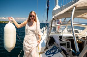 Blonde woman in white dress holding boat fender on sailboat deck with blue ocean and sky background.