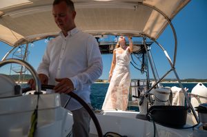 Bride and groom on a sailboat deck, man steering wheel, woman in wedding dress looking up, blue ocean background.