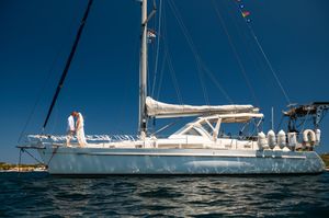 White sailboat anchored in calm blue waters under a clear sky, with a person standing on deck near the mast.