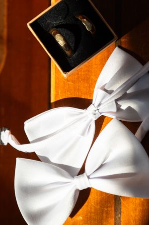 White bow ties displayed on wooden surface with gold wedding rings in black jewelry box.
