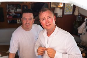 Two men in white shirts pose together indoors, one buttoning his shirt, with a decorated wall in the background.