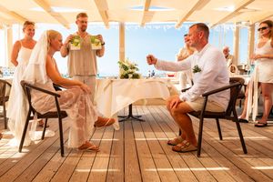 Couple toasting at an outdoor wedding reception on a wooden deck, with guests and a waiter serving in the background.