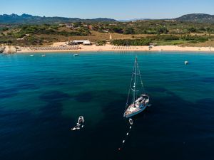 Aerial view of a sailboat anchored in turquoise waters near a sandy beach with lush hills in Sardinia, Italy.