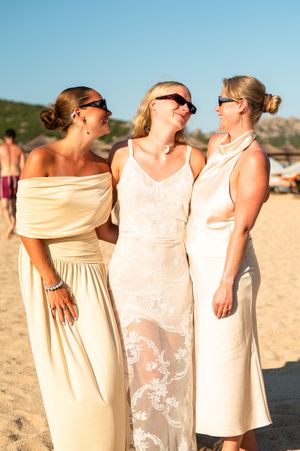 Three women in elegant cream dresses and sunglasses smiling together on a sunny beach wedding celebration.
