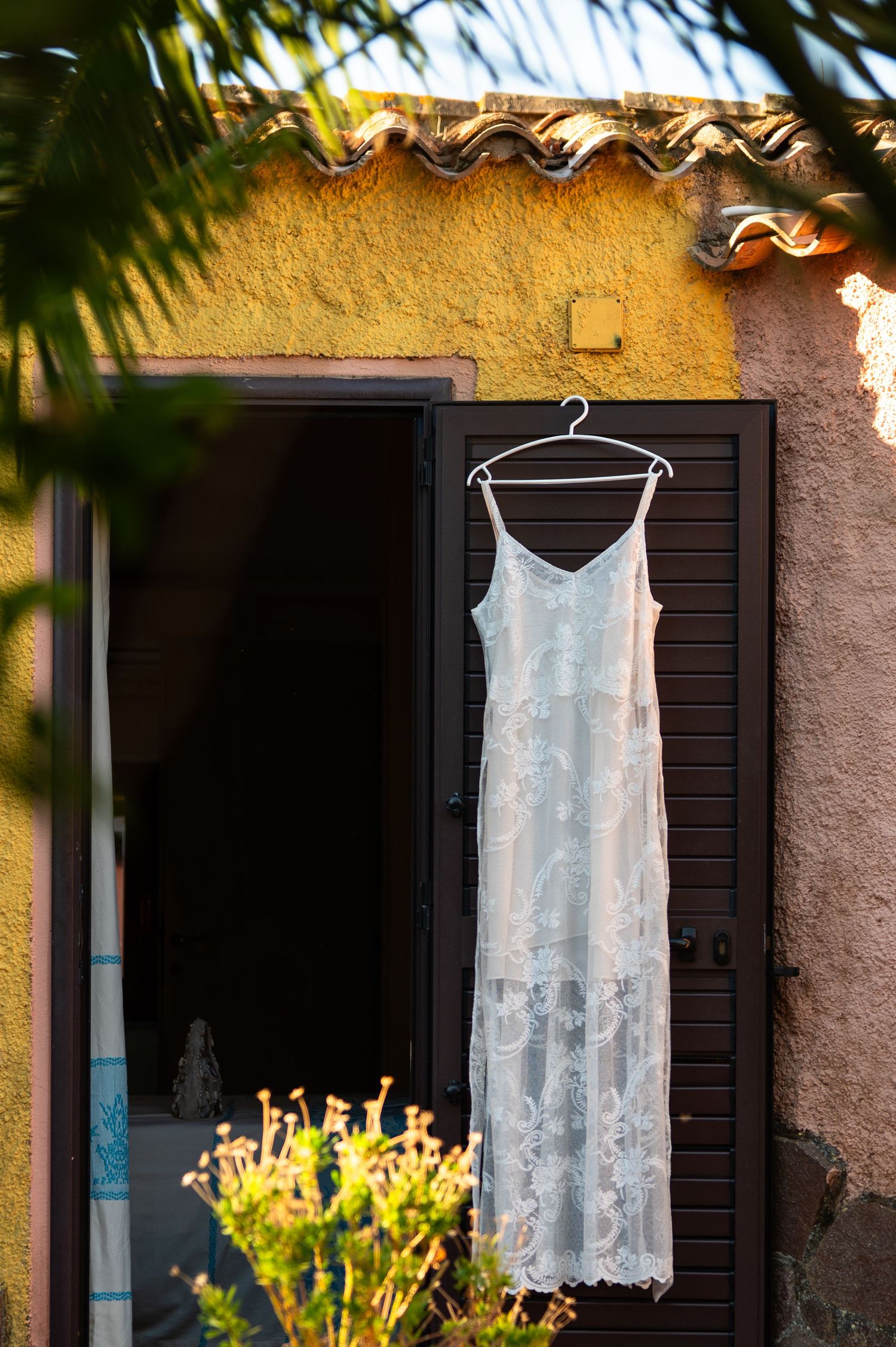 White lace wedding dress hanging on a hanger outside a rustic Mediterranean villa door in warm sunlight.