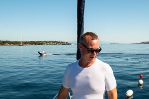 Man in white t-shirt and sunglasses on a boat with calm blue sea and sailboat in background.