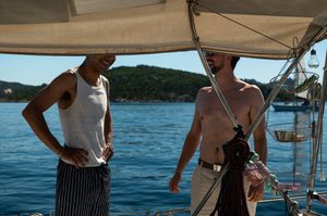 Two shirtless men standing on a sailboat deck with blue ocean and green hills in the background.