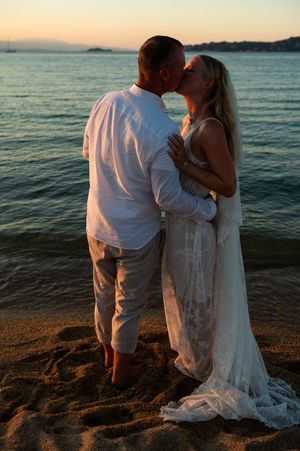 Couple sharing a romantic kiss on a sandy beach at sunset, dressed in white wedding attire by the ocean.