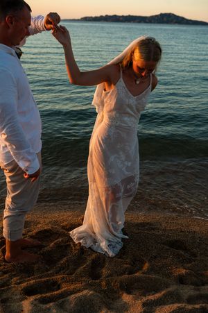 Bride in white lace gown dances with groom on sandy beach at golden sunset by calm ocean waters.