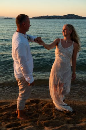 Couple in white attire holding hands on sandy beach at sunset with ocean and mountains behind them.