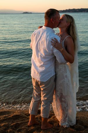 Couple sharing a romantic kiss on a sandy beach at sunset, bride in white lace gown, groom in white shirt.