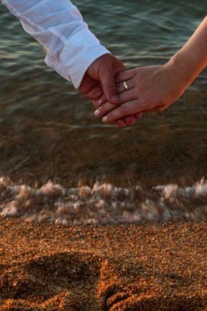Couple holding hands on a sandy beach at sunset, one wearing a wedding ring and white shirt.