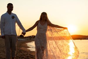 Bride and groom walk hand in hand on a beach at sunset, her lace veil flowing in the breeze.