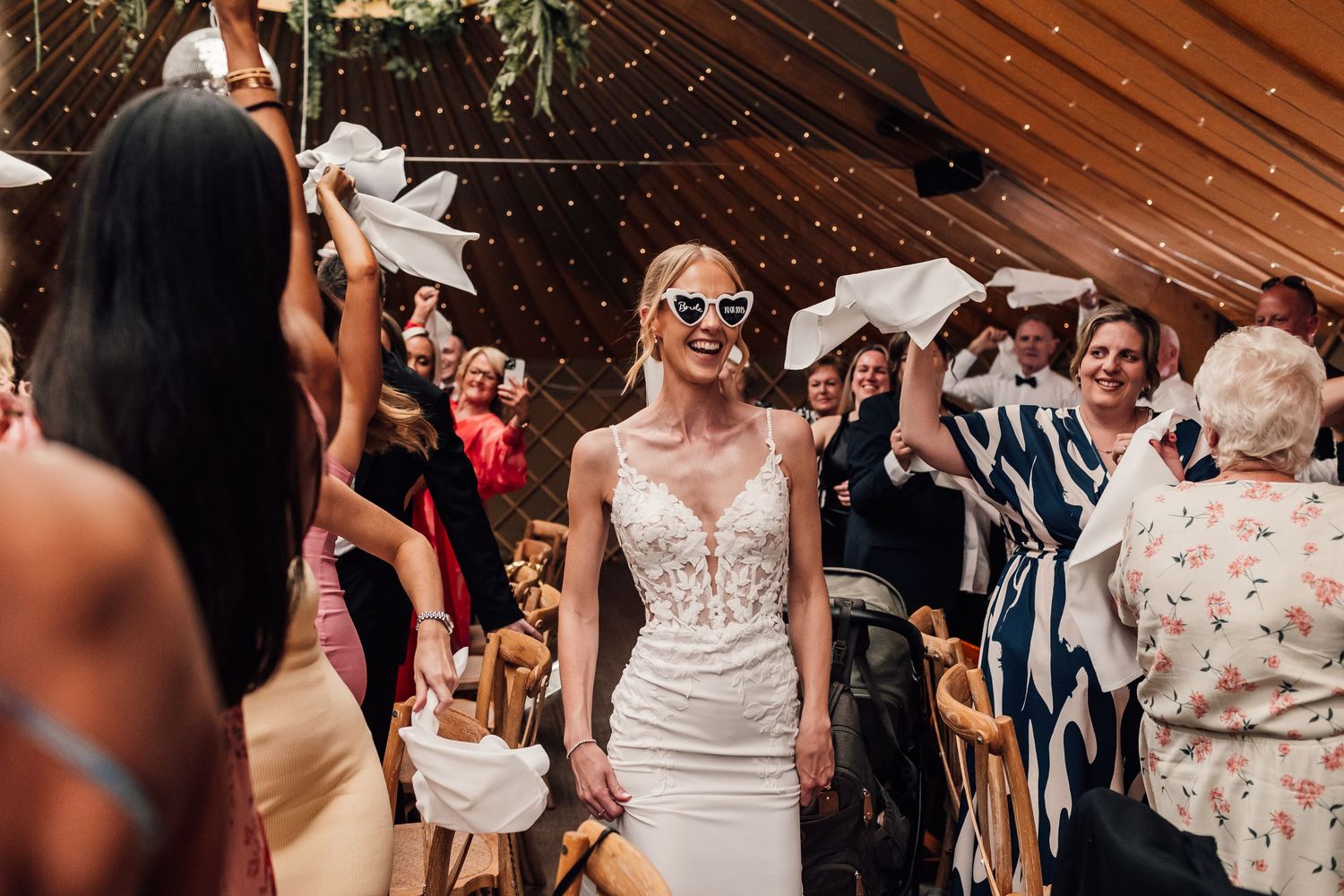 Wedding guests dance and celebrate on a rustic wooden dance floor under twinkling string lights at an indoor reception.