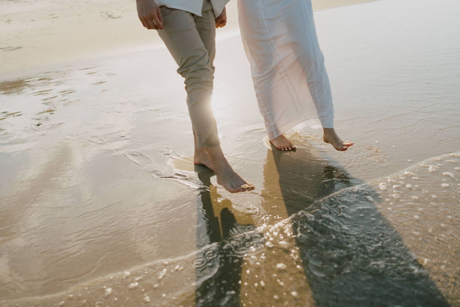 Two people standing barefoot in shallow ocean waves on a beach during golden hour.