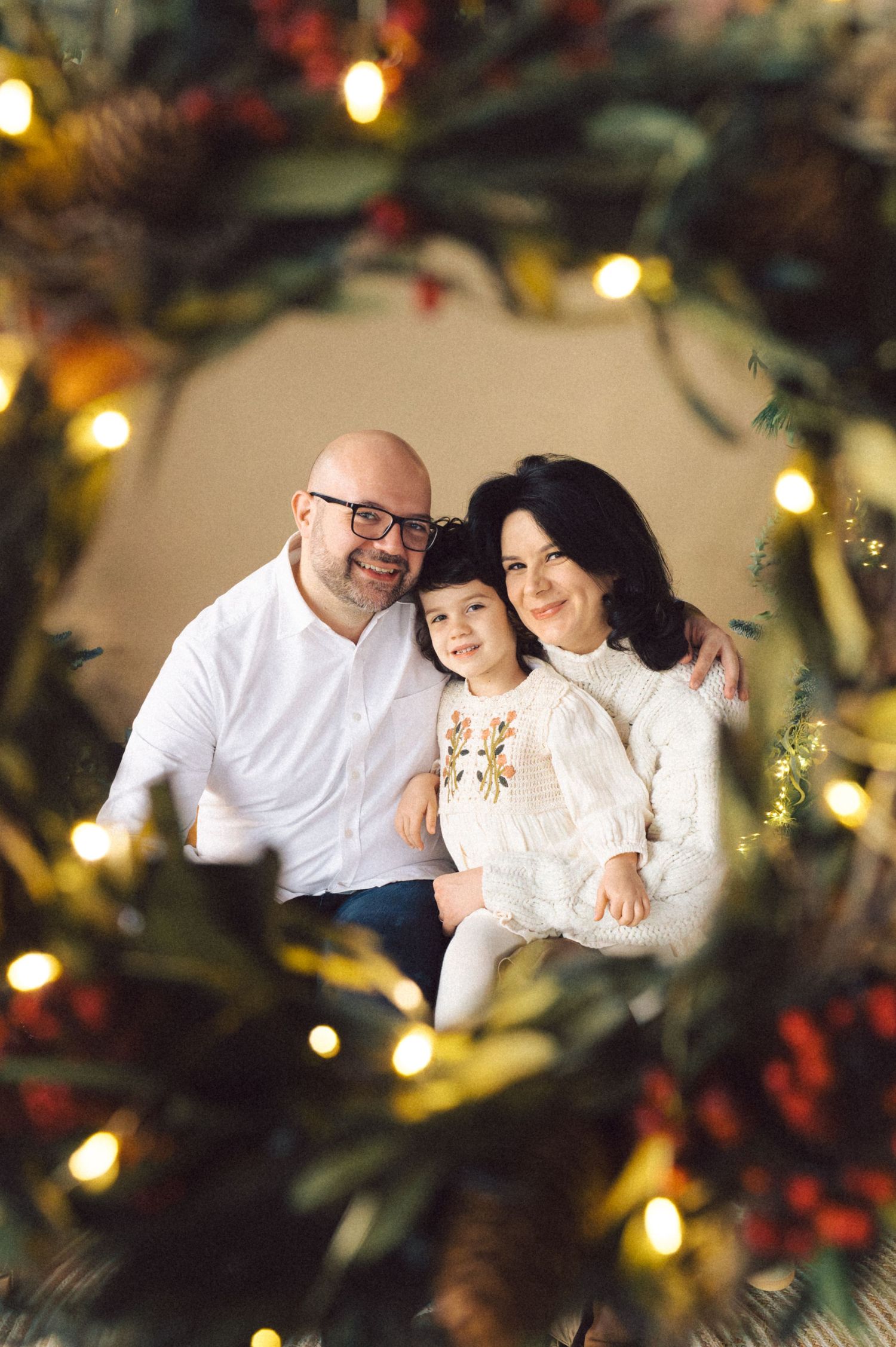 A family portrait taken through a Christmas wreath with holiday lights and decorations glowing in the foreground.