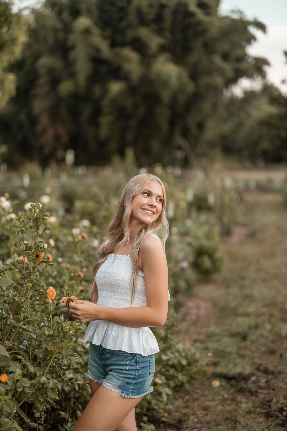 A person in denim shorts and white top posing playfully in a sunny flower field with plants and blooms.