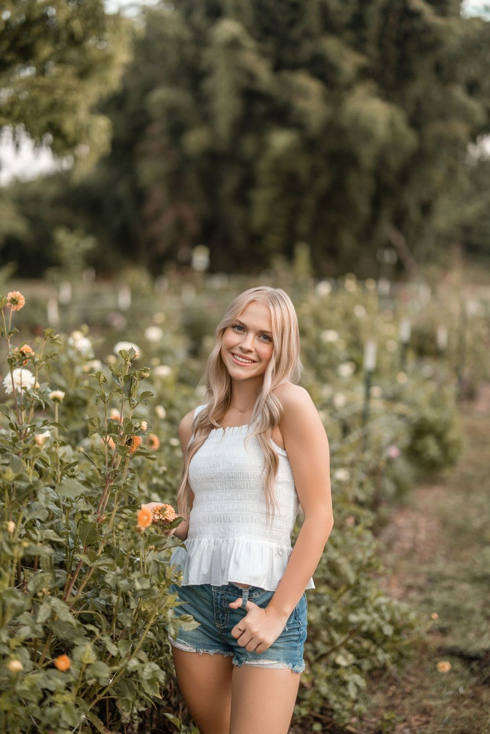 A person poses in a white top and denim shorts among blooming sunflowers in a garden on a sunny afternoon.